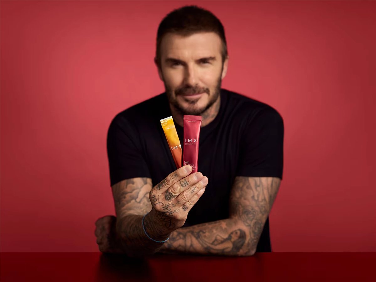 David Beckham holding two IM8 supplement sachets against a deep red backdrop.