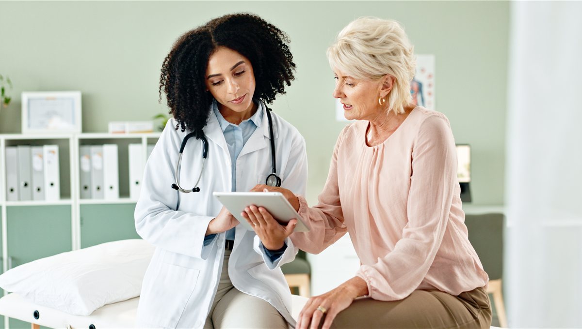 A doctor holding a tablet in conversation with an older patient in a consulting room.