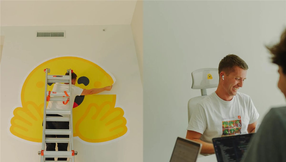 An office wall painted with the Hugging Face emoji, with someone working at a desk beside it.