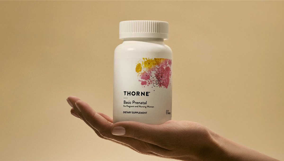 A hand holding a bottle of Thorne Basic Prenatal supplements against a sand-coloured background.