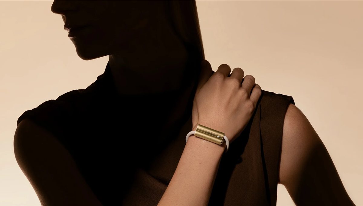 A hand resting on a shoulder, wearing a plain gold cuff that houses a health tracker.