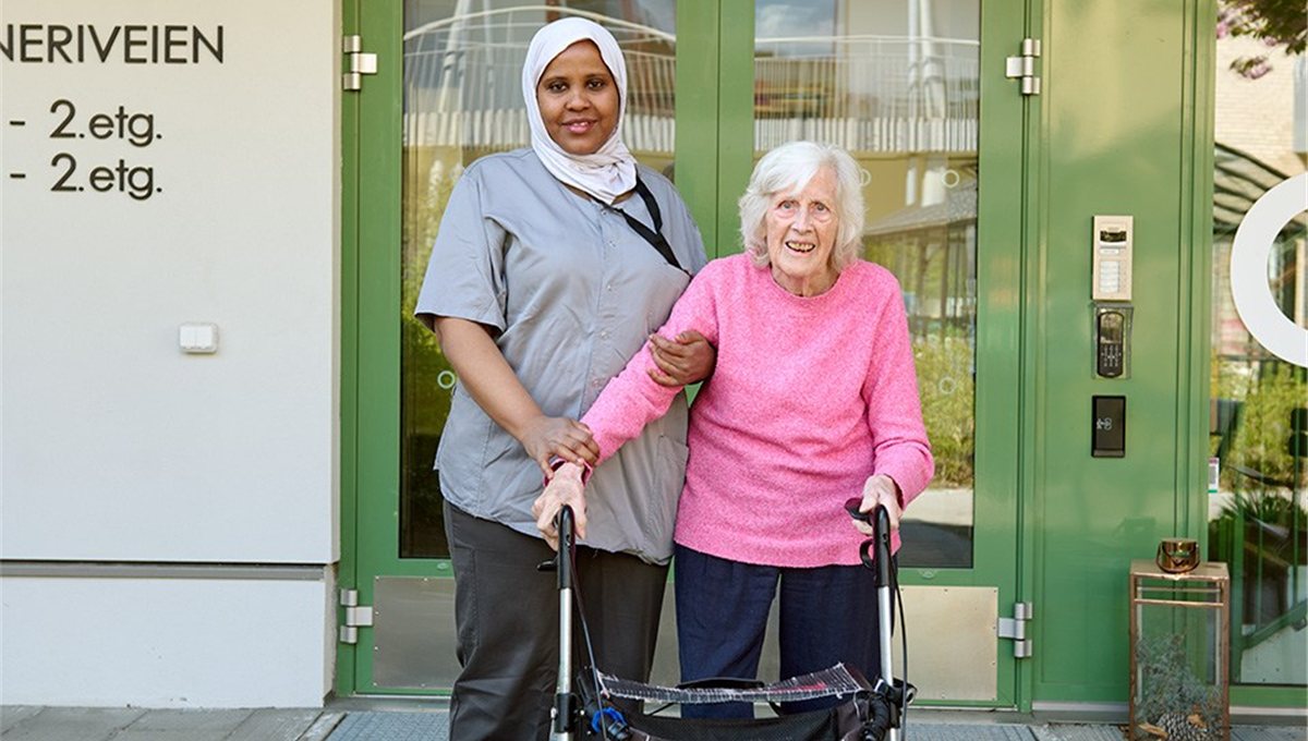 A care worker walking arm in arm with an elderly woman using a wheeled frame outside a care home.