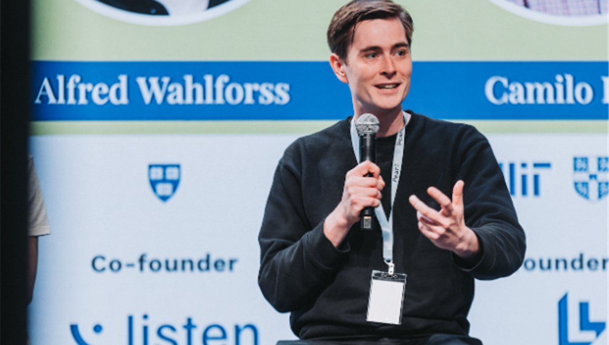 A founder speaking with a microphone on stage, his name on the screen behind him.