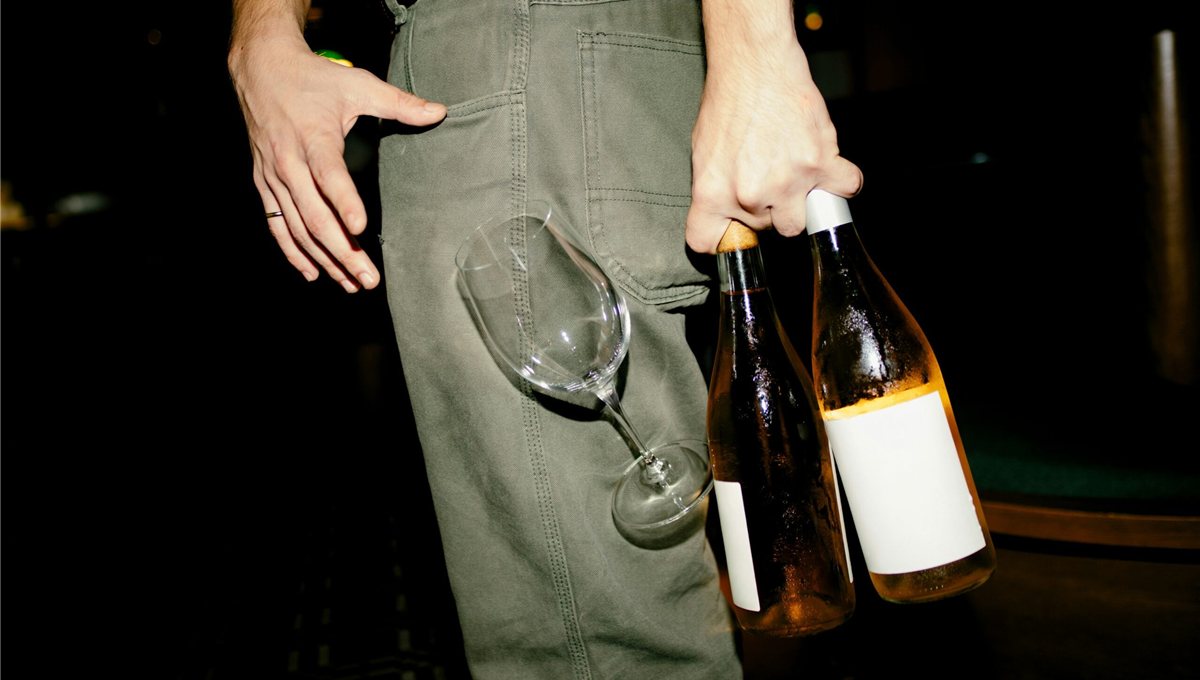 A person holding two wine glasses in one hand and two bottles of orange wine in the other.